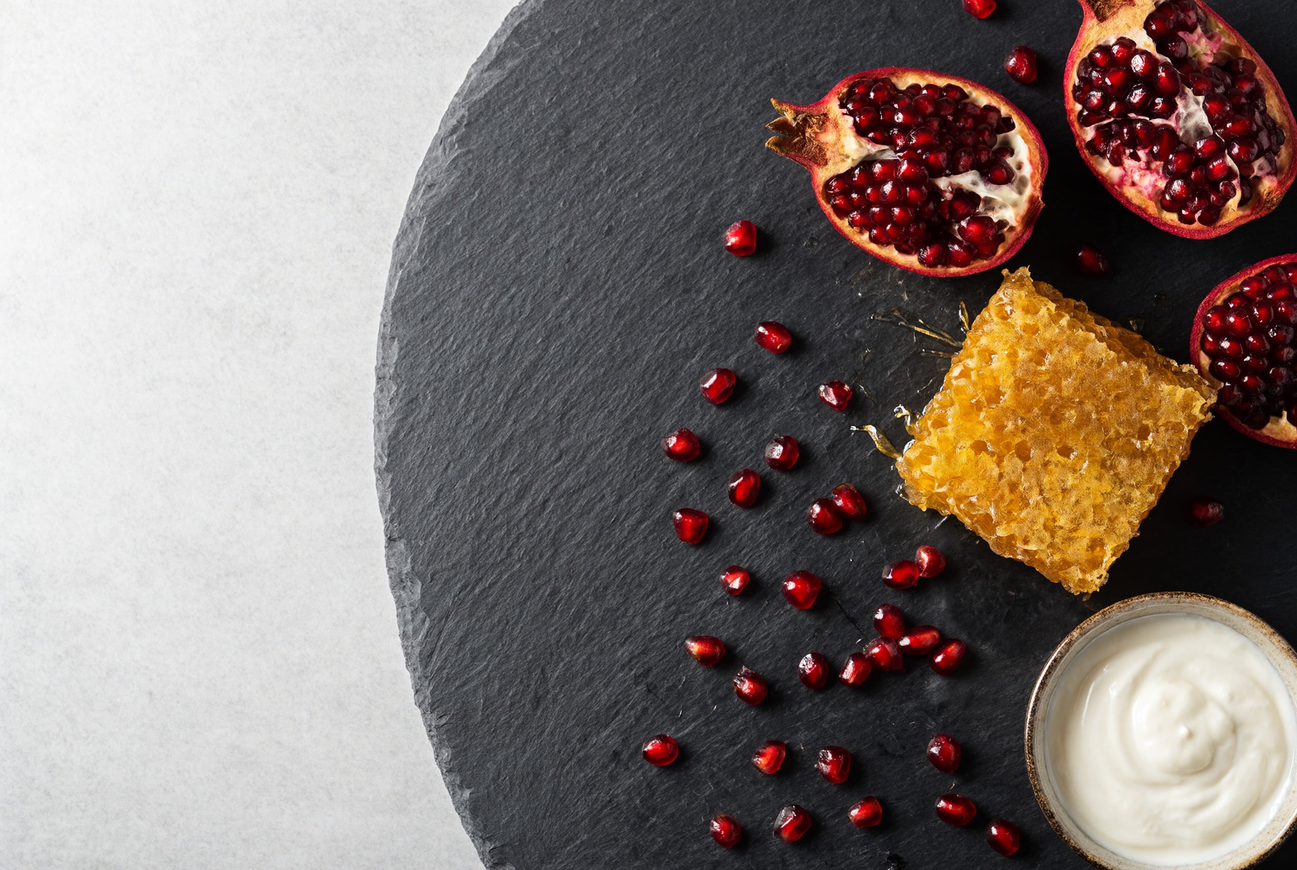 Overhead flat lay of pomegranate halves, ruby seeds, golden honeycomb and a bowl of yogurt on a dark slate board