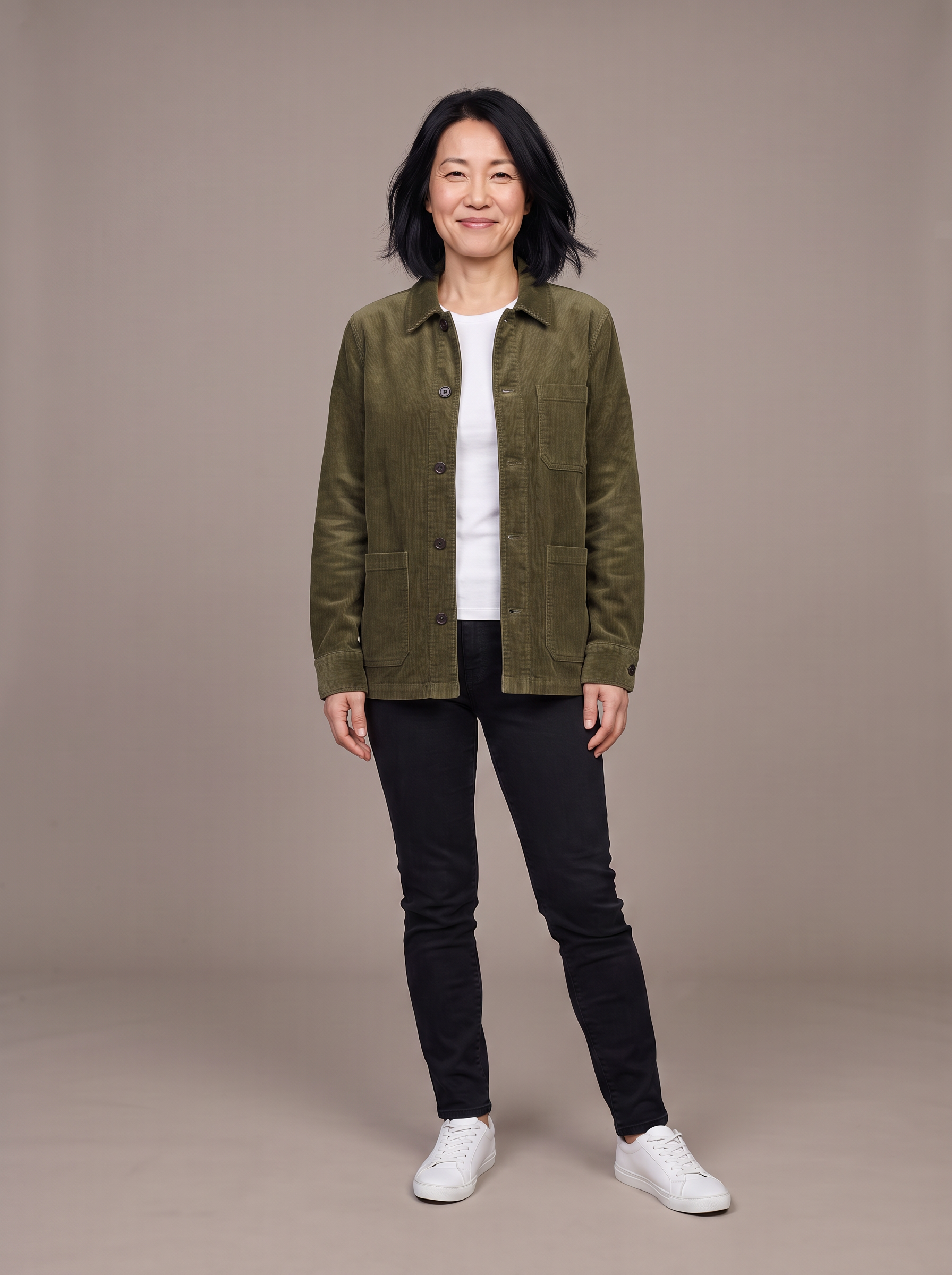 A petite woman in her forties with a black bob wears an olive corduroy chore jacket open over a white tee on a grey backdrop