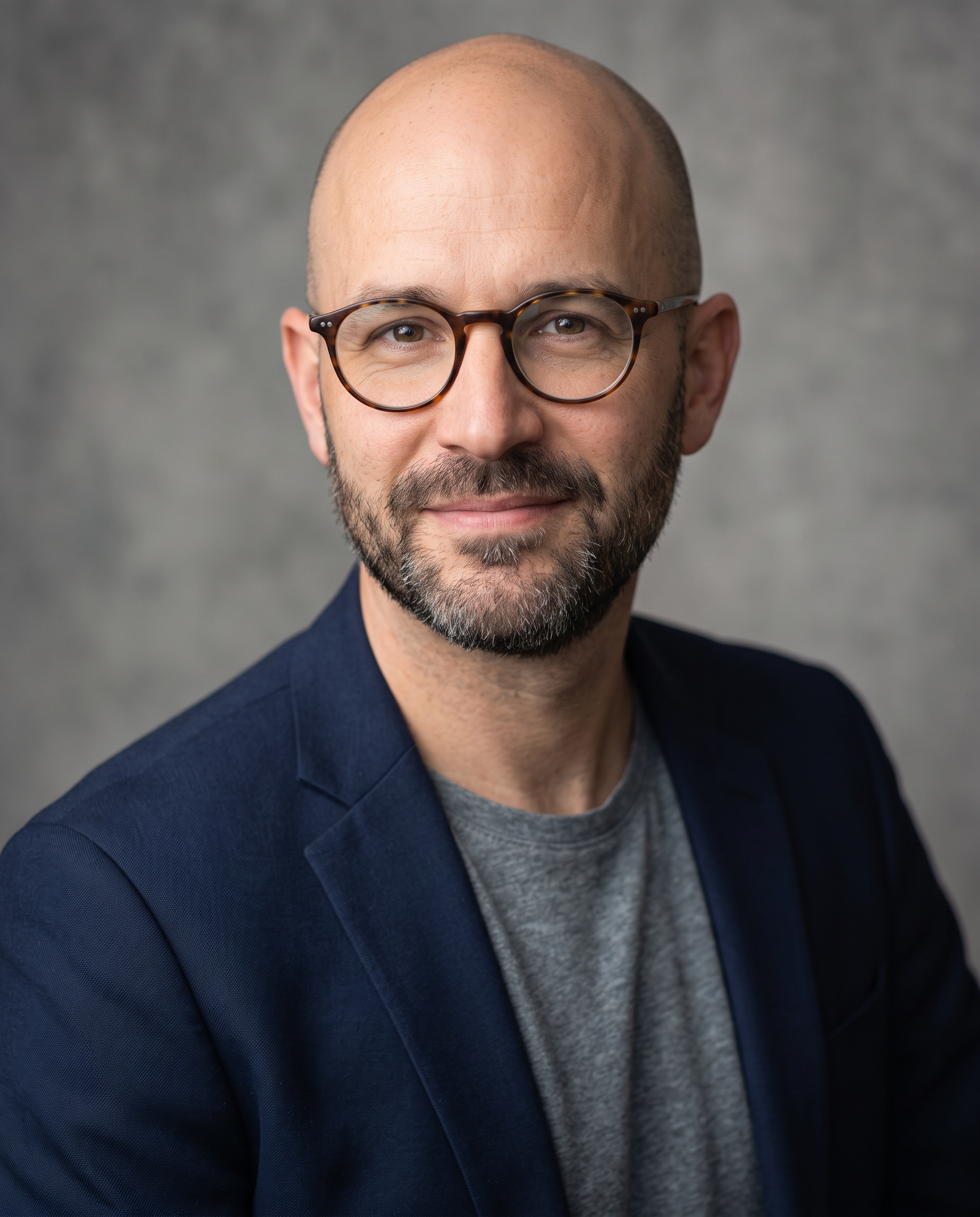 A bald man in his forties with round tortoiseshell glasses and a navy blazer smiles against a grey backdrop