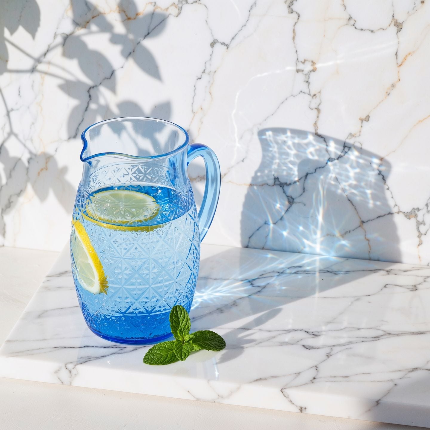 Cut glass blue pitcher of water with lemon slices on a white marble ledge, bright blue caustic light rippling across the marble wall, a sprig of mint and soft leaf shadows