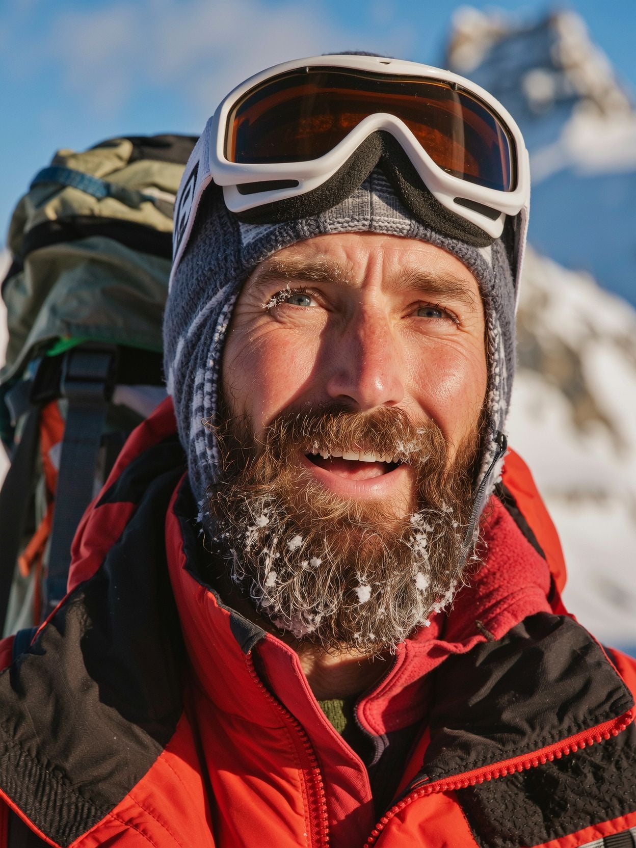 Close portrait of a mountaineer with a frosted brown beard, ice on his eyelashes, goggles on his knit hat and a red jacket, squinting into low sun with snowy peaks behind